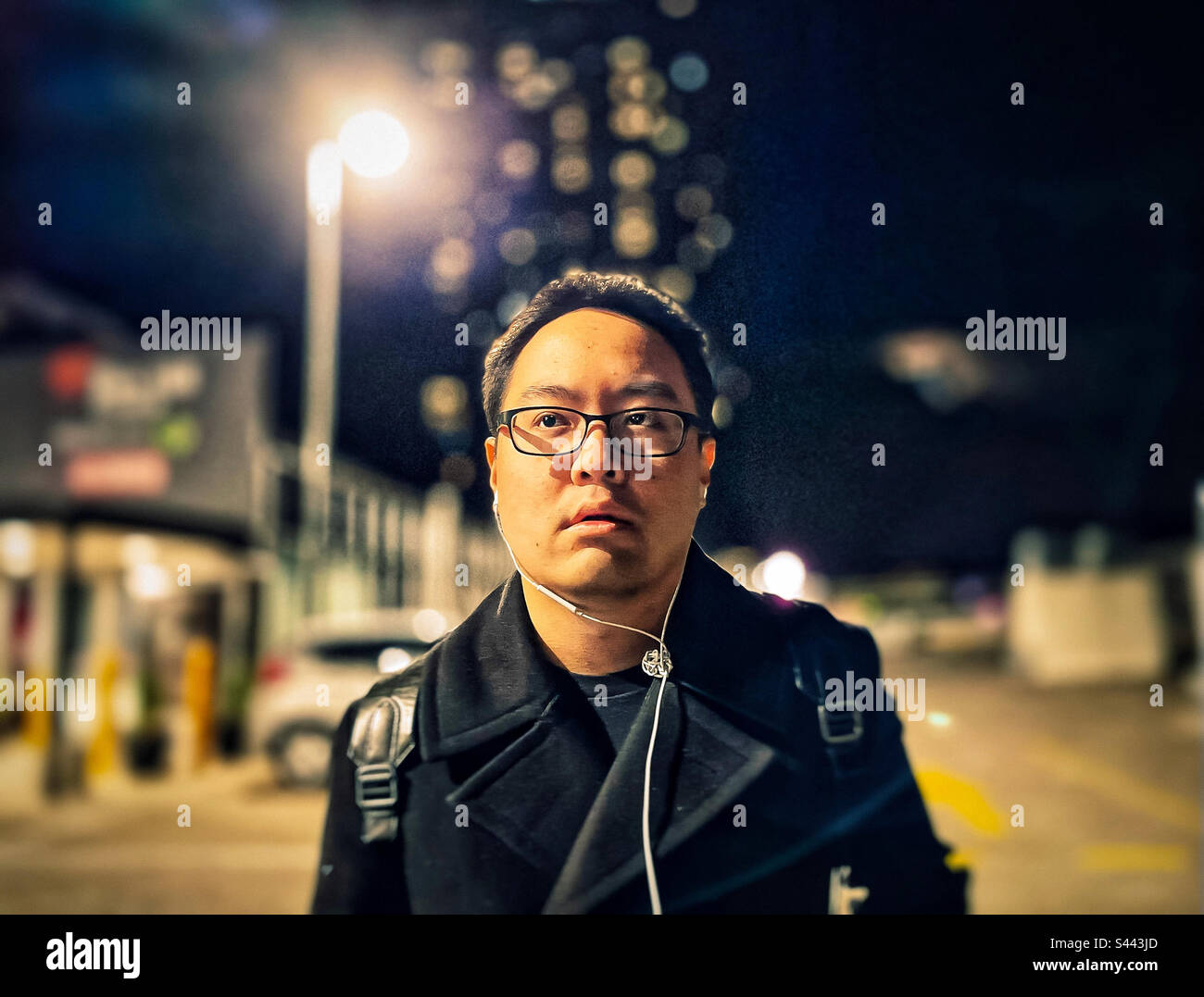 Ritratto di giovane asiatico in occhiali nel parcheggio di notte contro lampione, luci, costruzione e cielo notturno. Messa a fuoco in primo piano. Bokeh. Auricolari. Foto Stock