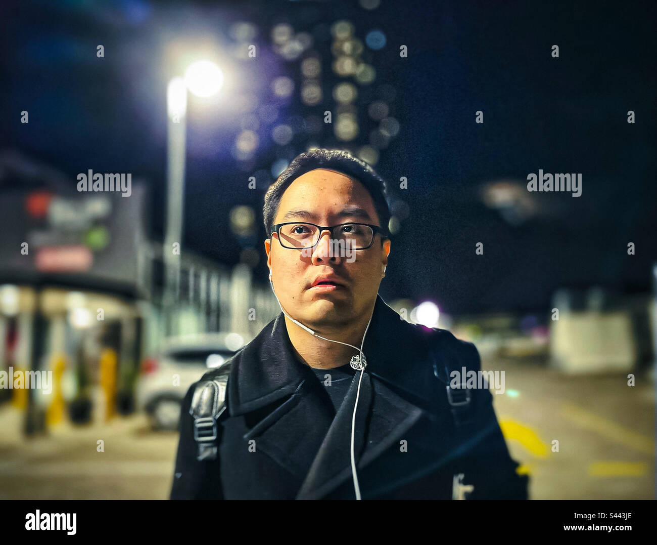 Ritratto di giovane asiatico in occhiali e auricolari nel parcheggio di notte contro lampione, luci di strada, edifici e cielo notturno. Messa a fuoco in primo piano. Bokeh. Foto Stock
