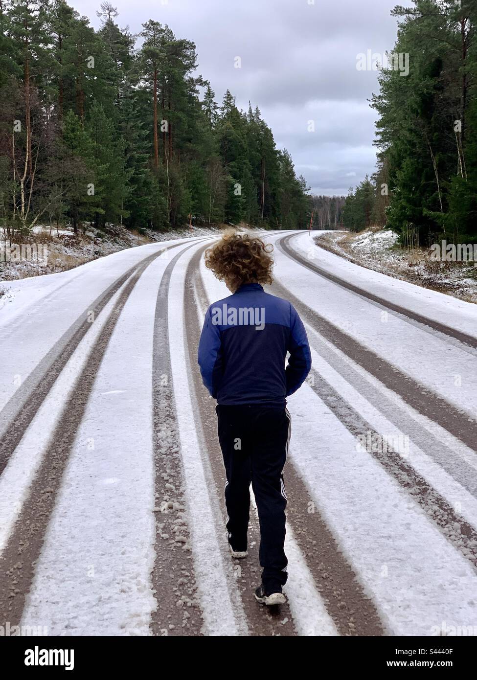 Un giovane adolescente che cammina da solo nel profondo del pensiero su una strada invernale coperta di neve in neve con le piste per pneumatici auto Foto Stock