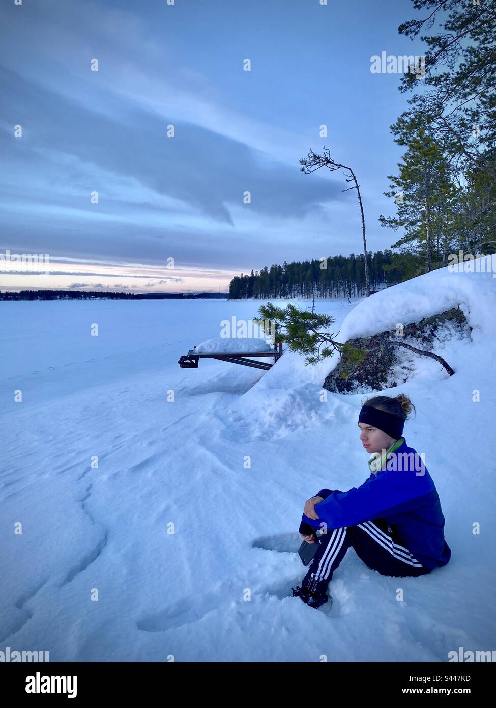 Un giovane adolescente seduto da solo sulle rive di un enorme lago ghiacciato coperto di neve nel profondo del pensiero in Finlandia Artico in inverno Foto Stock