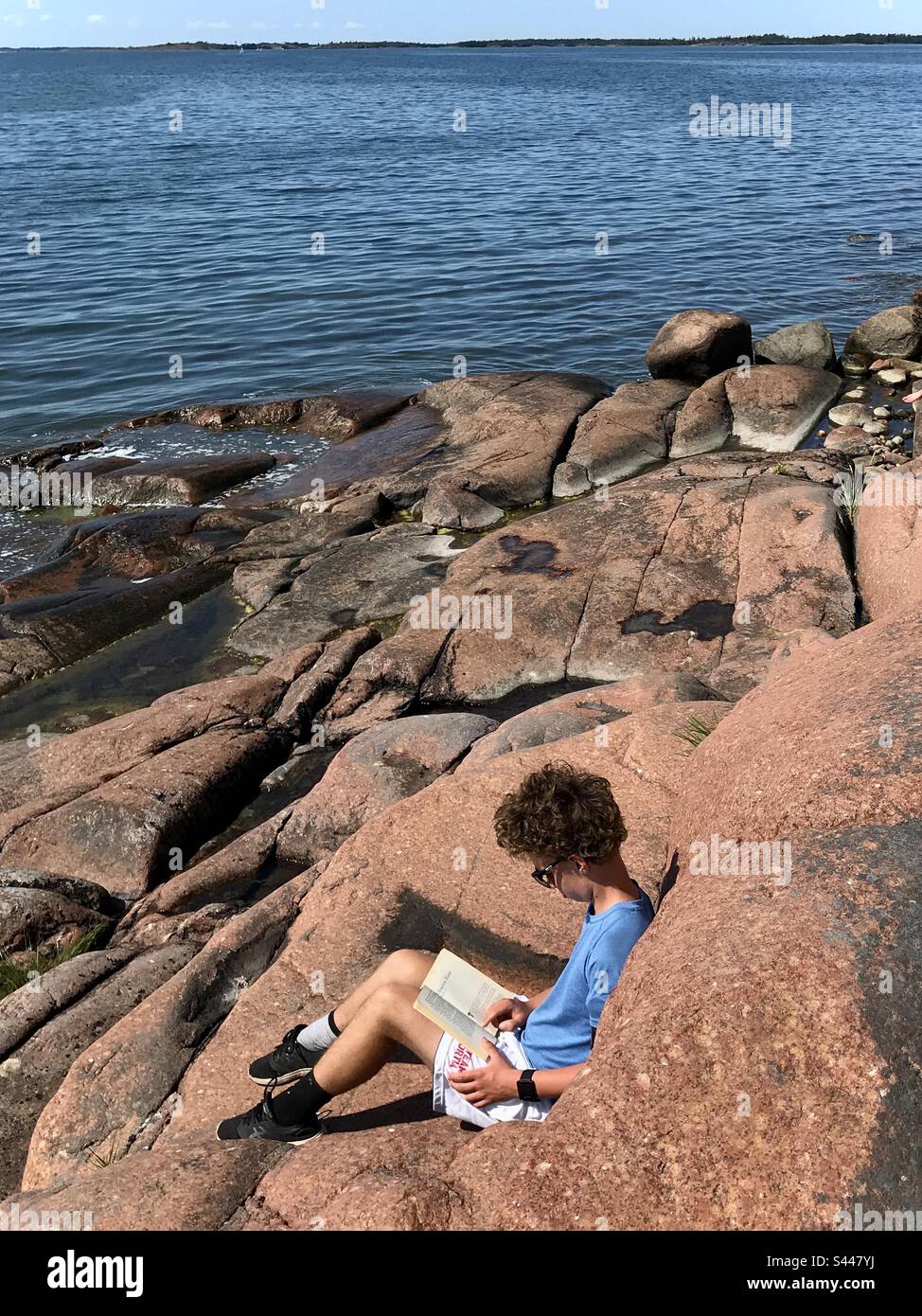 Un giovane ragazzo adolescente che legge da solo seduto su una spiaggia di costa rocciosa penisola in una bella giornata estiva vicino al mare oceano Foto Stock