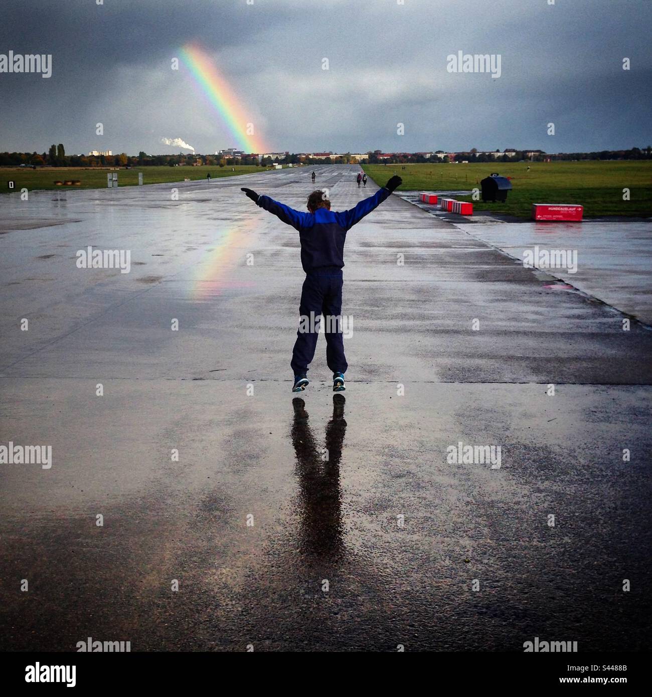 Un giovane ragazzo di 10 anni che salta fingendo di volare e atterrare sulla pista sotto un arcobaleno all'aeroporto Tempelhof di Berlino, il campo aereo del centro città in tempo di guerra, che è ora un popolare parco pubblico Foto Stock