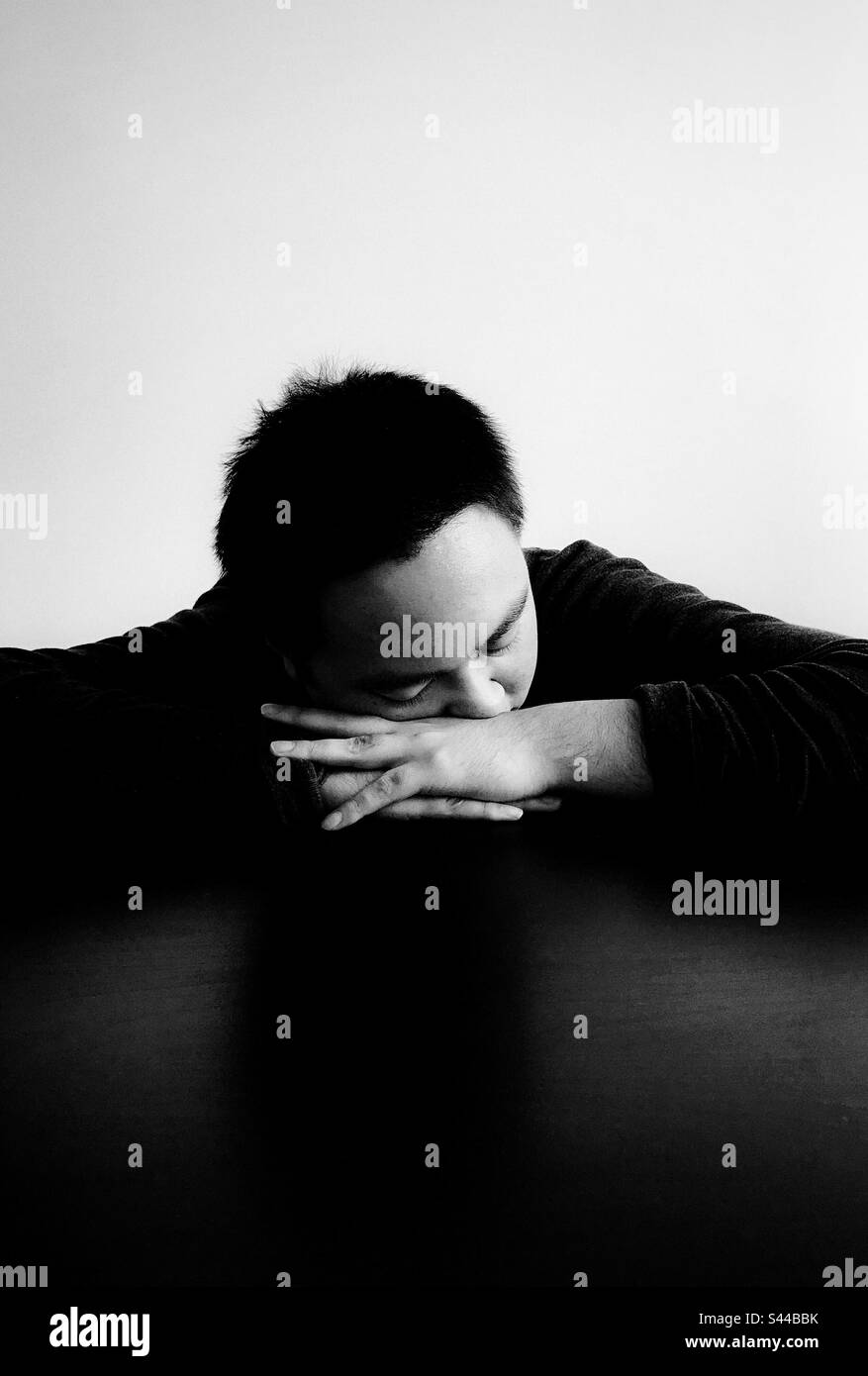Primo piano ritratto di un giovane asiatico con gli occhi chiusi e la testa appoggiata sulle mani ripiegate sul tavolo. Monocromatico. Spazio di copia. Oscurità. Emozione. Tristezza. Foto Stock Primo piano ritratto di un giovane asiatico con gli occhi chiusi e la testa appoggiata sulle mani ripiegate sul tavolo. Monocromatico. Spazio di copia. Oscurità. Emozione. Tristezza. Foto Stock