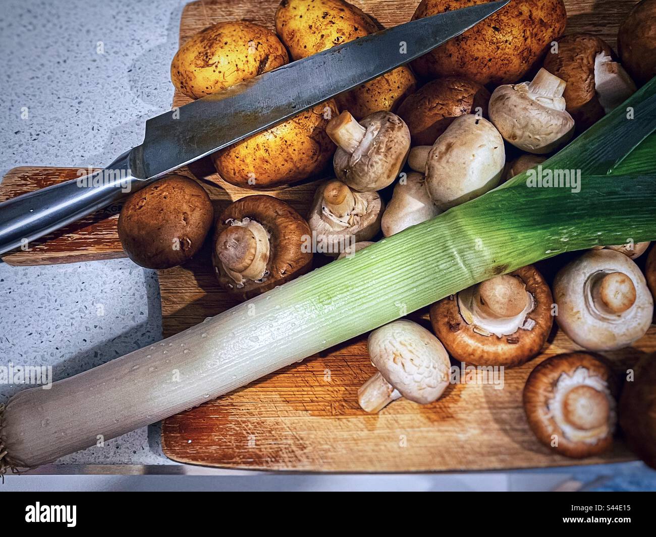 Ingredienti per crema di funghi, porri e zuppa di patate. Vista ad angolo alto di funghi bottoni marroni e bianchi, un gambo di porro e patate crema olandesi sul tagliere di legno insieme a un coltello. - Immagine stock catturata con smartphone