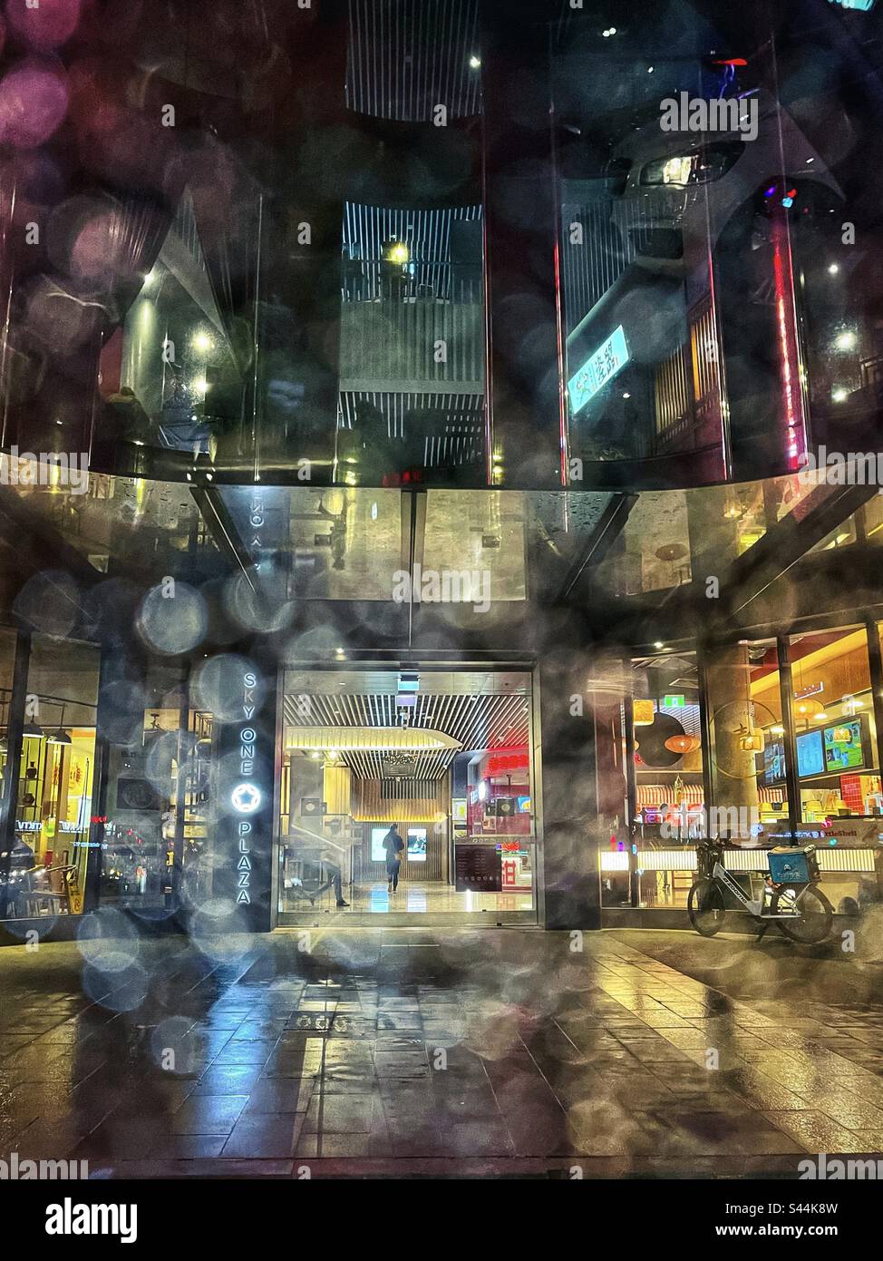 Vista attraverso le finestre di vetro bagnato del marciapiede e del quartiere dei ristoranti all'interno della facciata di vetro illuminata dell'edificio in una notte piovosa a Box Hill, Victoria, Australia. Foto Stock