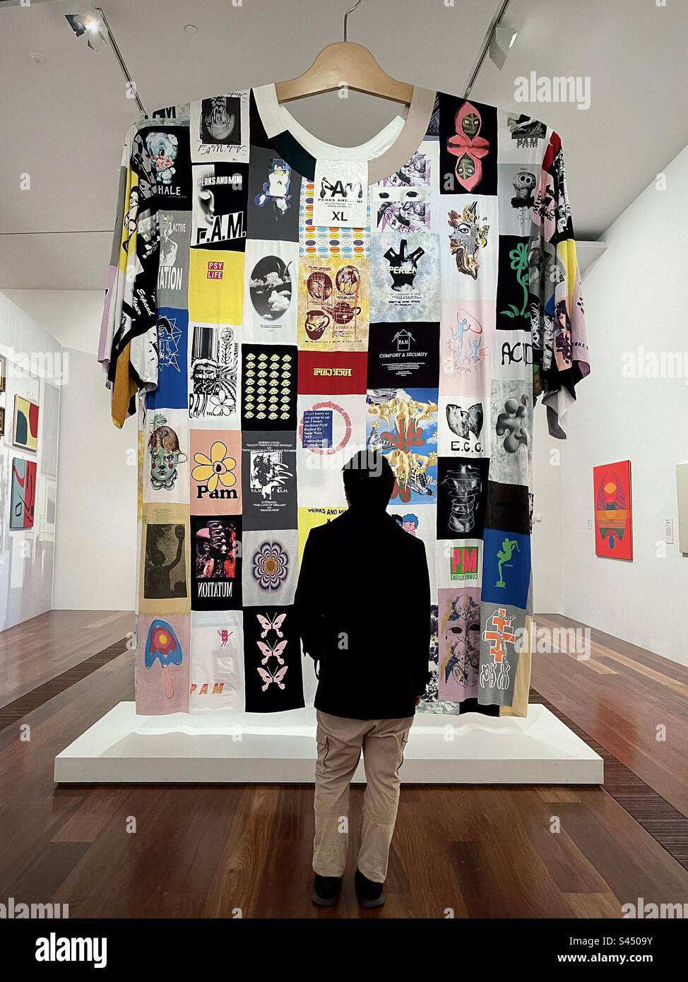 Vista posteriore dell'uomo che guarda una maglietta gigante, autoritratto con T-shirt, di Perks e Mini in mostra alla mostra Melbourne Now allo Ian Potter Centre: NGV Australia, Melbourne, marzo-agosto, 2023. - Immagine stock catturata con smartphone