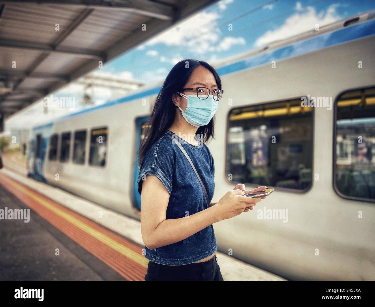 Giovane donna asiatica con occhiali e maschera viso che usa il telefono cellulare alla stazione ferroviaria. - Immagine stock catturata con smartphone