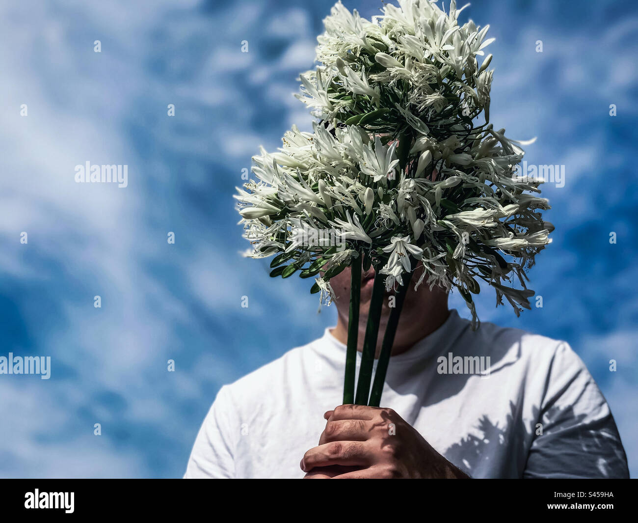 Ritratto di un giovane uomo in T-shirt bianca con un bouquet di fiori bianchi di Agapanthus africanus, detto giglio africano, contro il nuvoloso cielo blu. Faccia oscurata. Moda. Tema primaverile. - Immagine stock catturata con smartphone