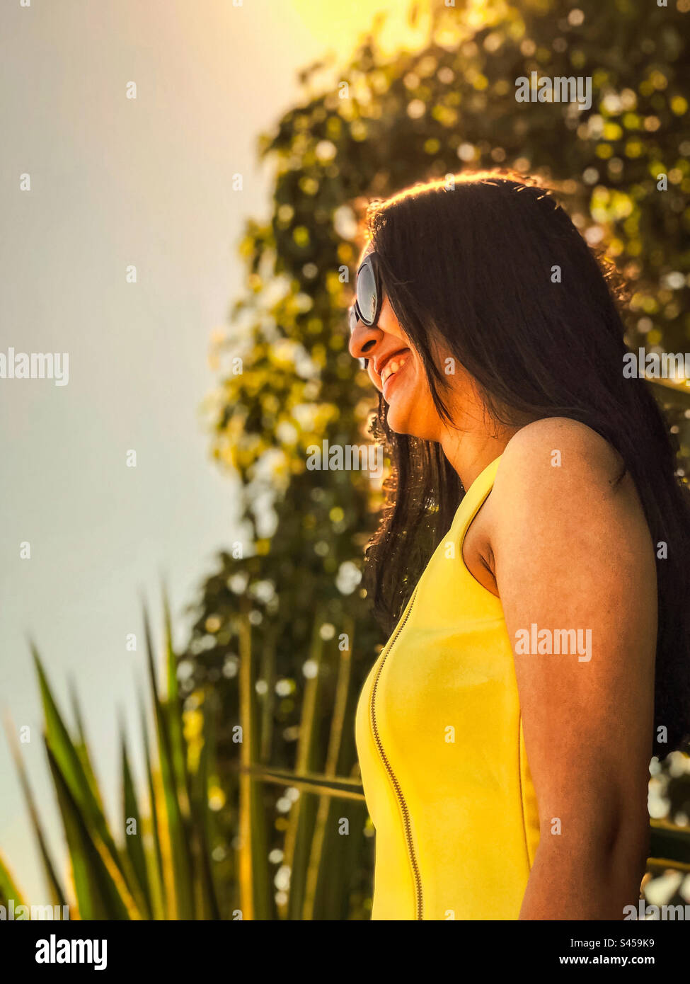 Ritratto di una giovane donna sorridente con occhiali da sole e vestito giallo contro piante e alberi, bagnata dalla luce del sole dorata. Moda. Felicità. - Immagine stock catturata con smartphone