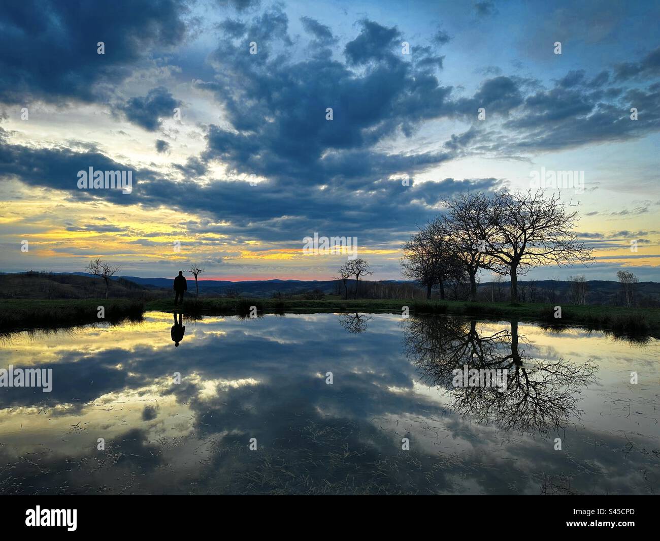 Nuvole e silhouette di un uomo che si riflette nell'acqua di un piccolo lago al tramonto Foto Stock