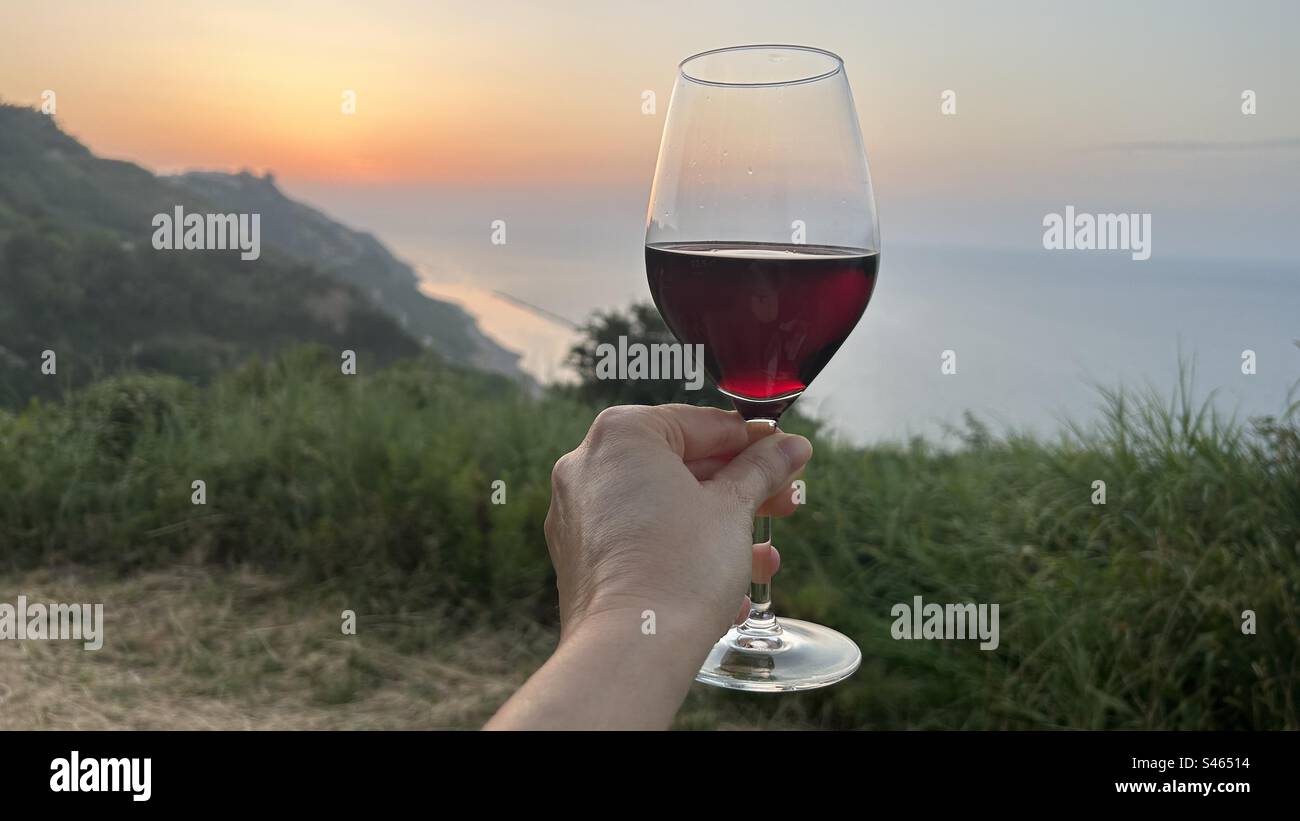 In mano un bicchiere di vino rosso esteso su un tramonto a picco sul mare nel parco naturale del Monte San Bartolo Foto Stock