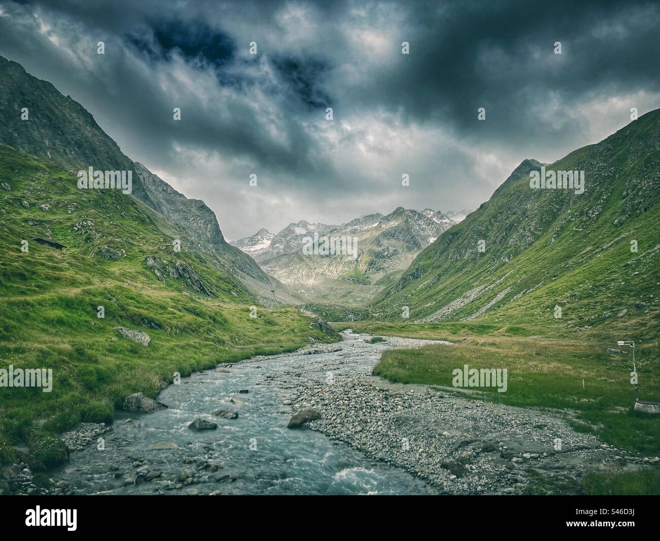 Torrente glaciale in una valle fiancheggiata da prati alpini di fronte con nuvole spettacolari e alte montagne sullo sfondo Foto Stock