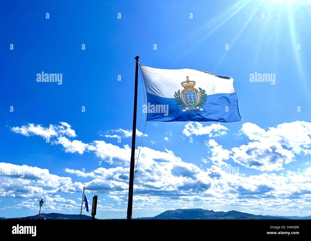 La bandiera della Repubblica di San Marion, con le sue bande azzurre e bianche e il loro stemma nazionale al centro, ondeggia contro il cielo blu con nuvole e raggi di sole Foto Stock