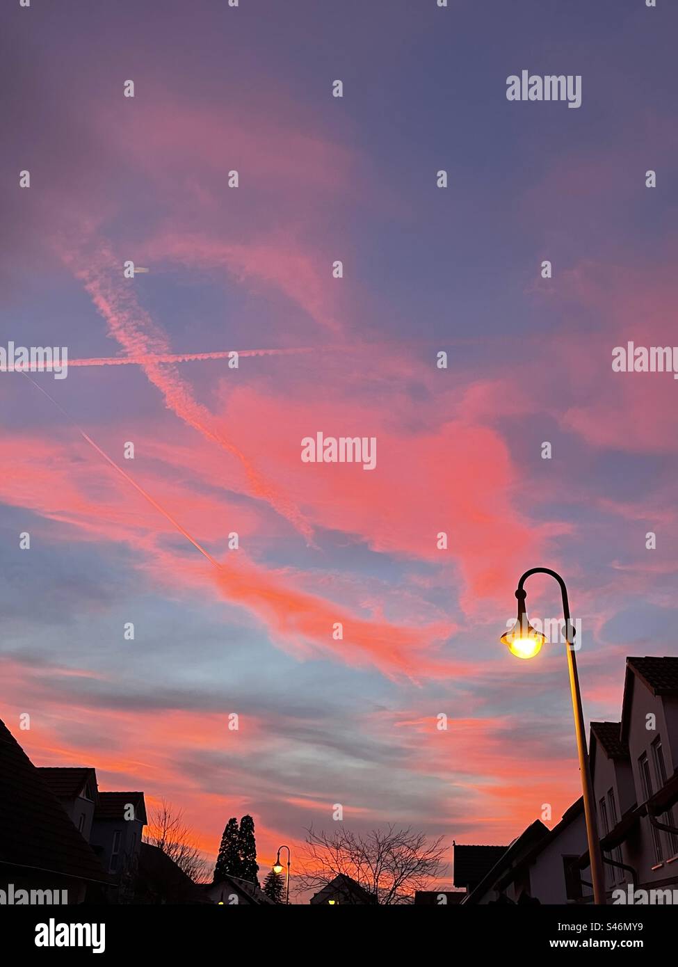 Sotto le sfumature rossicce di un tramonto, un lampione emana il suo caldo bagliore, segnando il passaggio dal giorno alla notte in una tranquilla scena urbana. Foto Stock