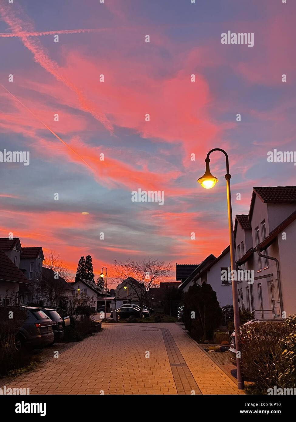 Sotto le sfumature rossicce di un tramonto, un lampione emana il suo caldo bagliore, segnando il passaggio dal giorno alla notte in una tranquilla scena urbana. - Immagine stock catturata con smartphone