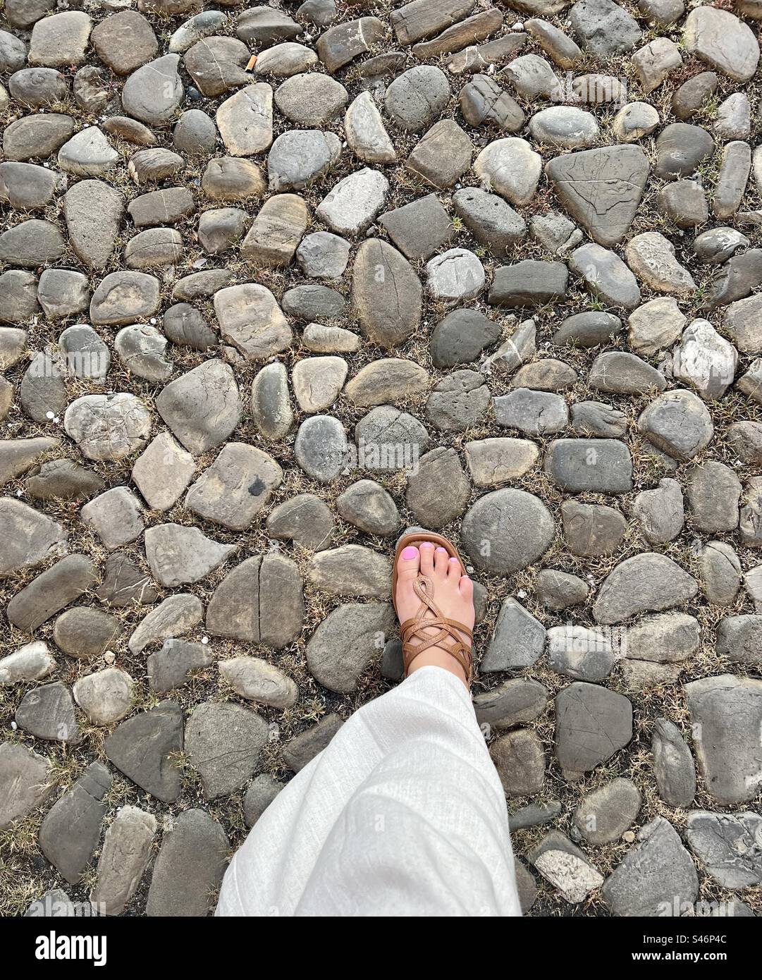 Piede femminile con pedicure rosa, che cammina su una strada acciottolata, con pantaloni di lino e un sandel abbronzato Foto Stock