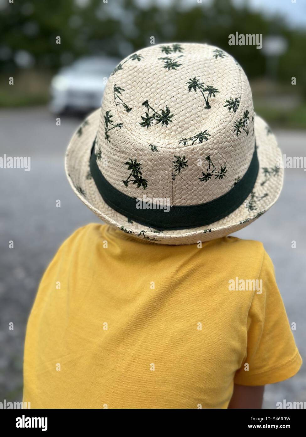 Un bambino con una t-shirt gialla siede e cappello con motivi di palma, rivolto verso l'esterno, osserva curiosamente l'auto davanti, catturando un momento sincero e intrigante. - Immagine stock catturata con smartphone