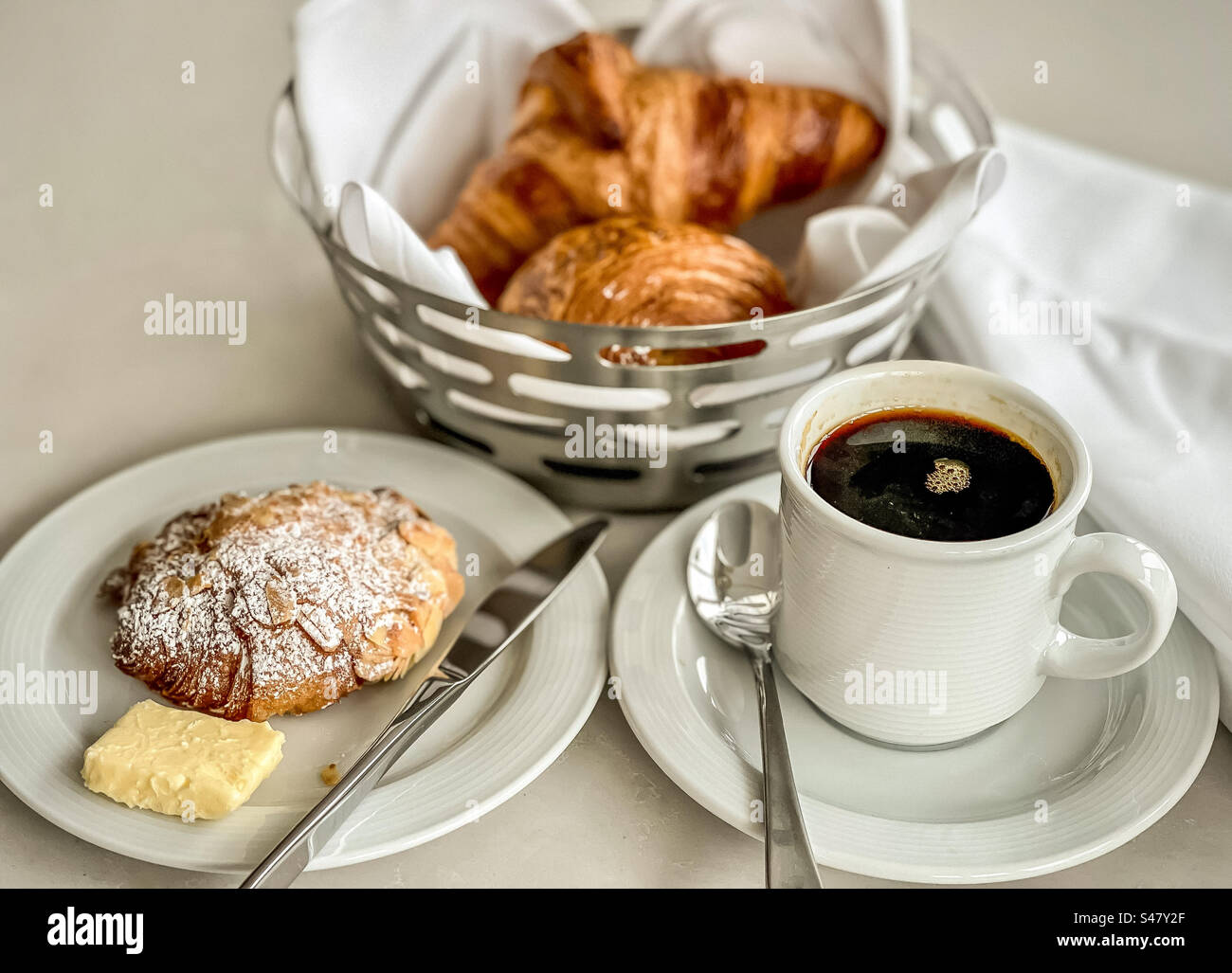 Vista dall'alto della colazione continentale: Croissant in ciotola metallica, pasticceria e burro su piatto con coltello e una tazza di caffè nero caldo. - Immagine stock catturata con smartphone