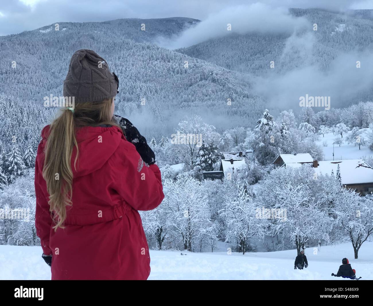 Una bambina vestita con un anorak rosso e un cappello di lana osserva un paesaggio di montagna innevato. In lontananza, altri due bambini scivolano sulla neve. Foto Stock