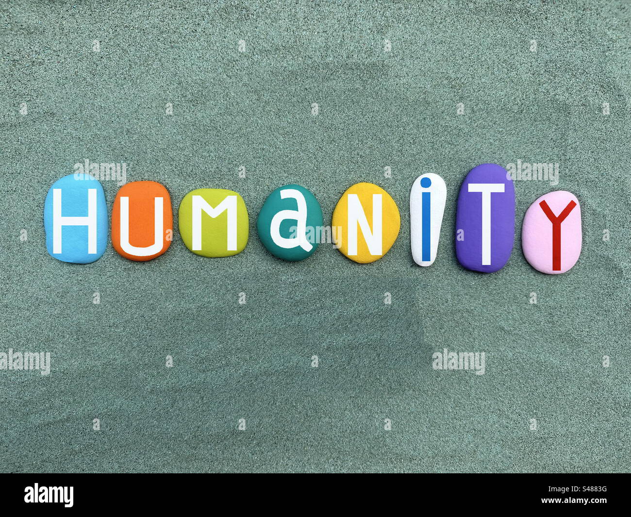 Parola umanitaria, slogan creativo o tipo di logo composto da lettere in pietra dipinte a mano multicolore su sabbia verde - Immagine stock catturata con smartphone
