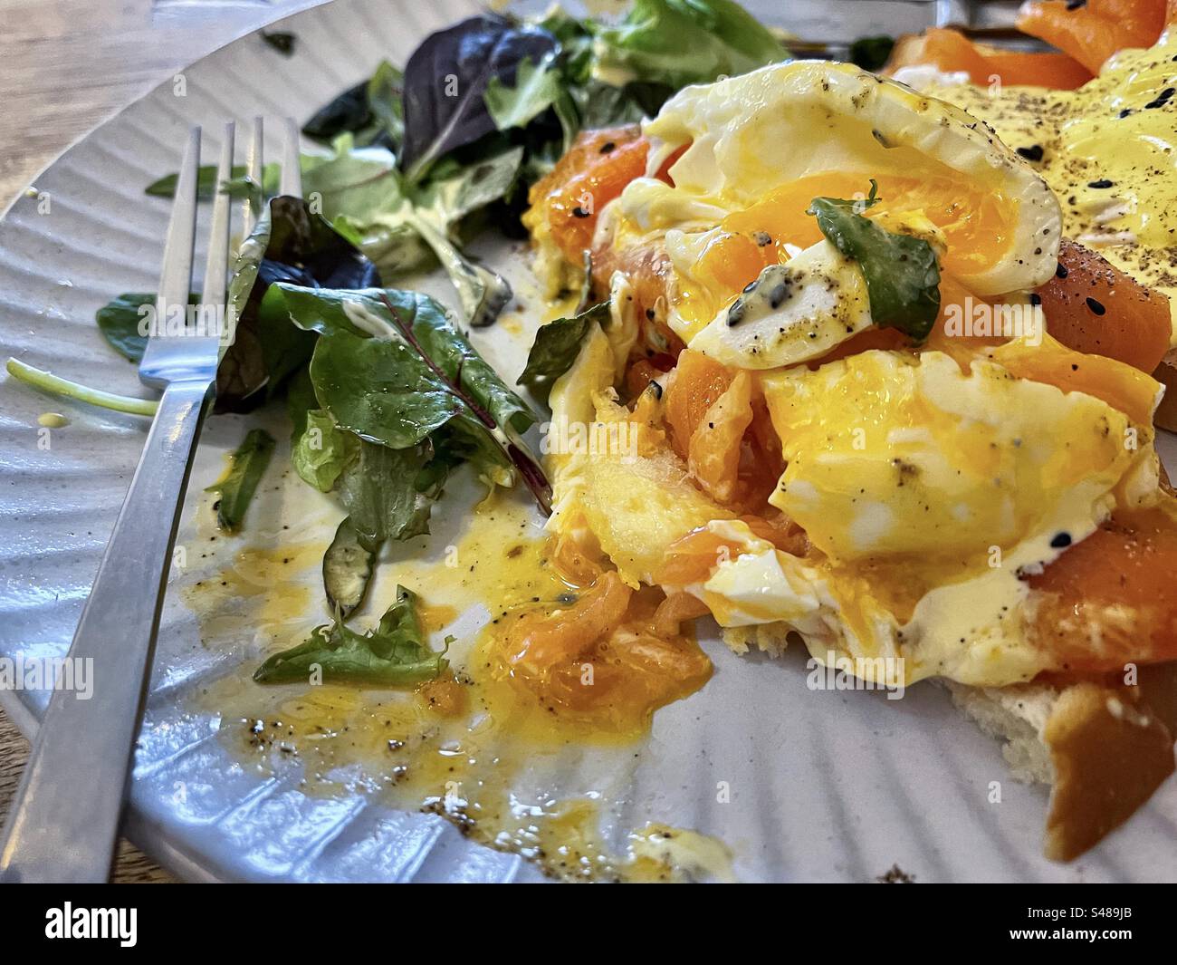 Primo piano di uova Benedict parzialmente consumate con salmone affumicato, salsa hollandaise e insalata di foglie di spinaci su piatto a tavola. - Immagine stock catturata con smartphone