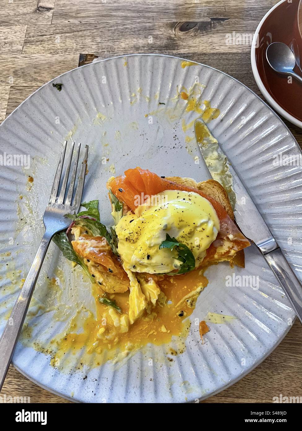 Direttamente sopra le uova Benedict parzialmente consumate con salmone affumicato, salsa hollandaise e foglia di spinaci su shokupan/pane al latte giapponese su piatto con posate sul tavolo. Foto Stock