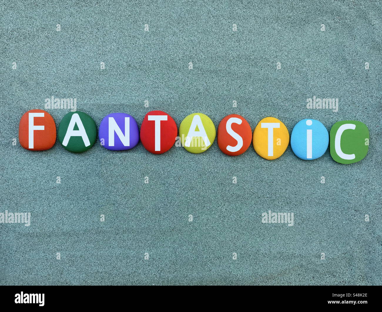 Parola fantastica composta da lettere di pietra dipinte a mano multicolore su sabbia verde - Immagine stock catturata con smartphone