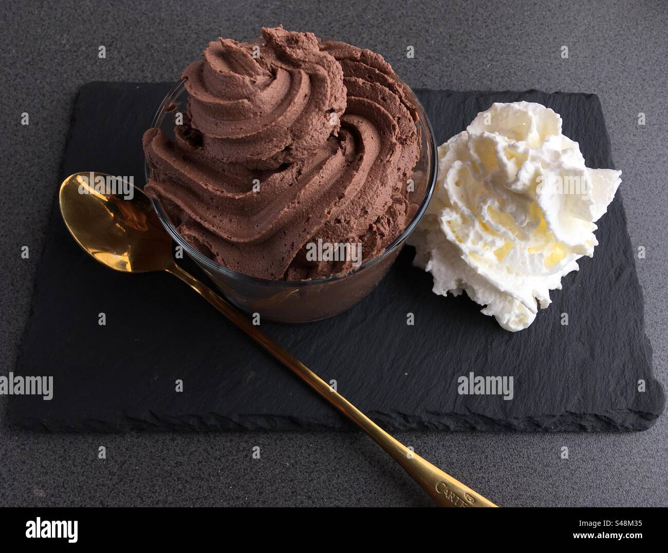 Primo piano di un dessert con mousse al cioccolato con panna montata su un piatto piatto piatto di pietra nera con un lungo cucchiaio d'argento Foto Stock