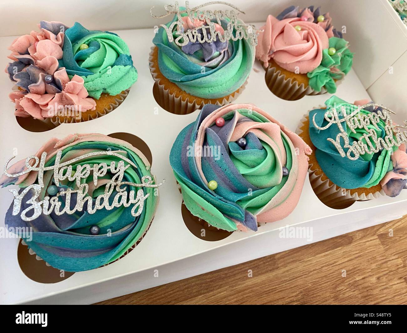 Una fotografia di 6 cupcake di compleanno in scatola e splendidamente decorati. Glassa turchese, viola e rosa, con decorazioni in argento per «buon compleanno». Foto Stock