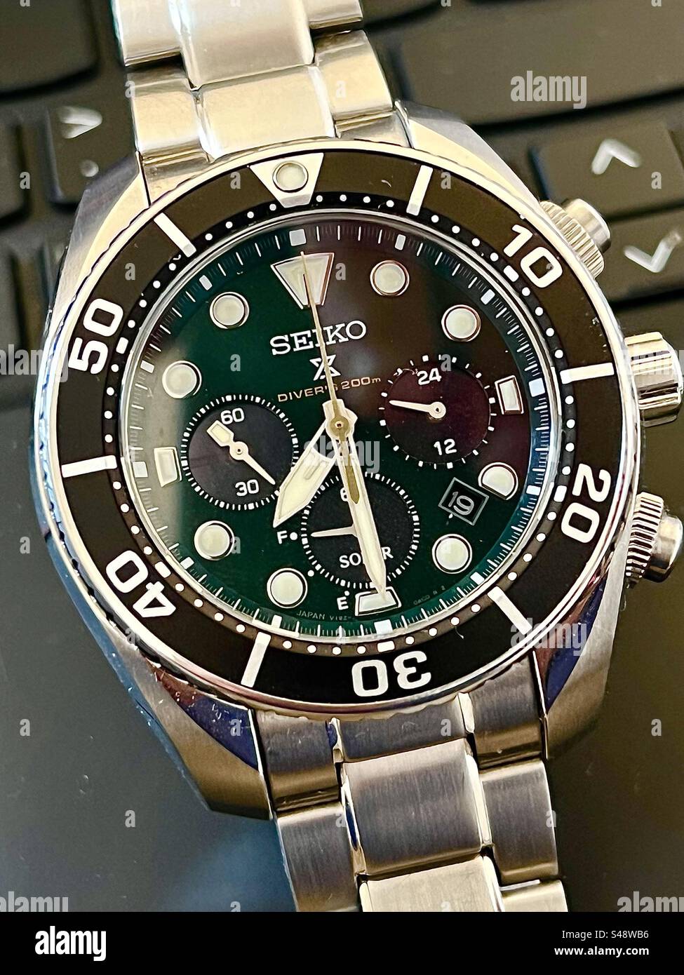 Primo piano di un orologio britannico con quadrante verde in edizione speciale Seiko Solar Chronograph Diver su uno sfondo nero con tastiera. - Immagine stock catturata con smartphone