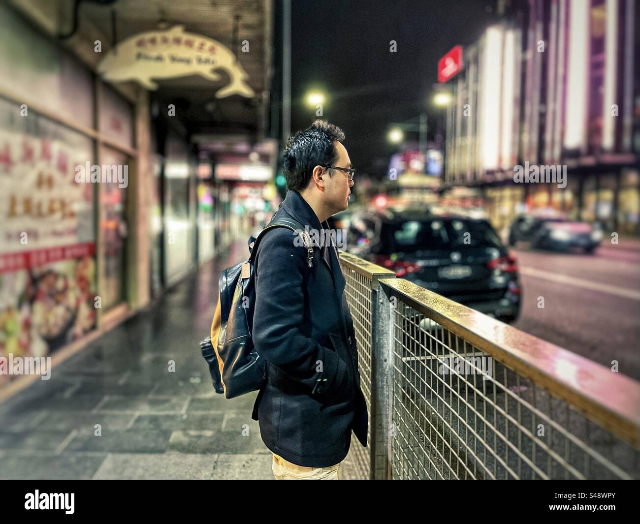 Vista laterale di un giovane asiatico in piedi sul marciapiede accanto alla trafficata strada della città di notte, di fronte agli edifici illuminati e ai negozi. Concentrati sul primo piano. Foto Stock