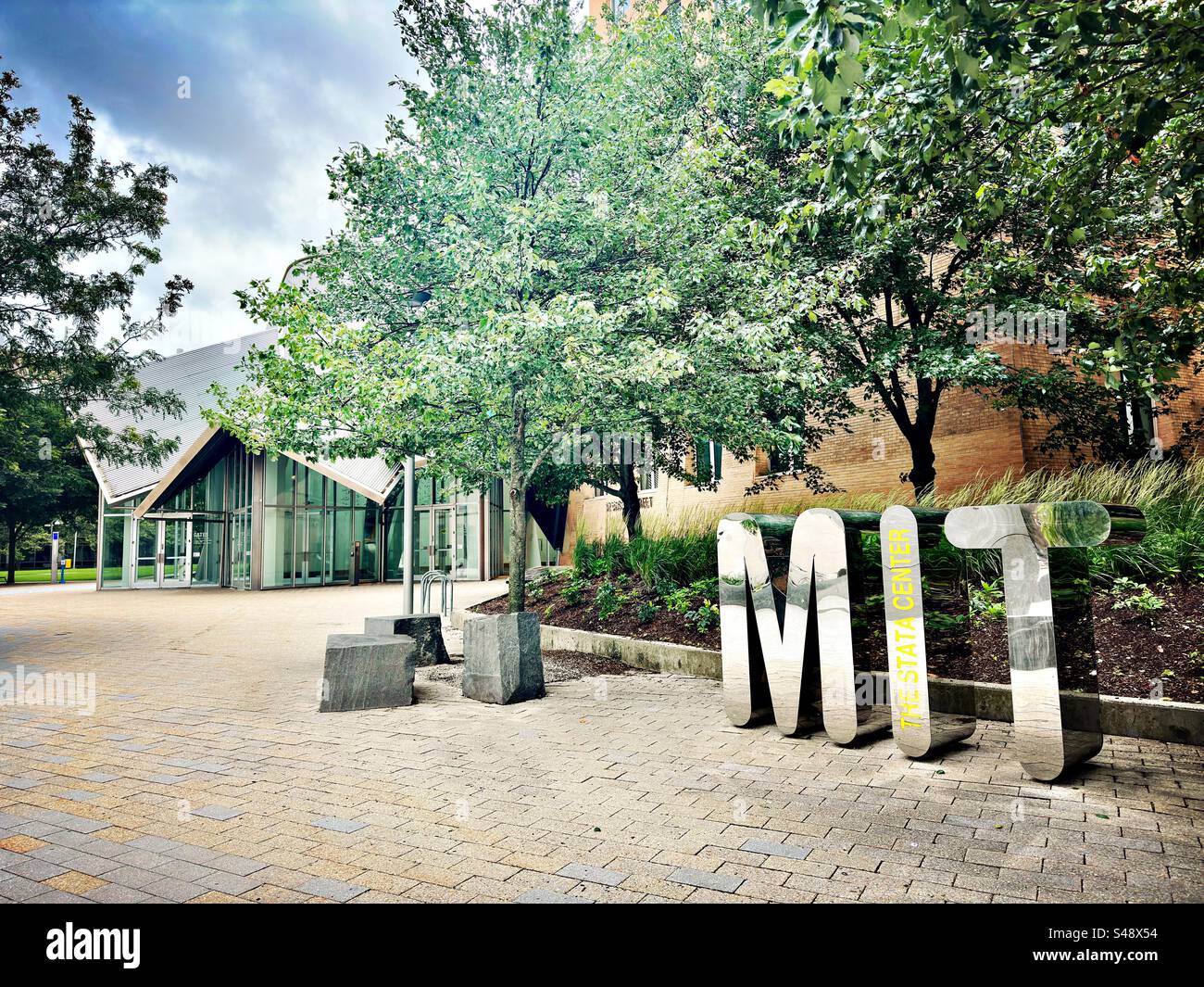 Cambridge, Massachusetts, USA: Massachusetts Institute of Technology - The statata Center. Insegna moderna e brillante per il MIT sul marciapiede. Edifici e alberi come parte della scena universitaria del campus. - Immagine stock catturata con smartphone