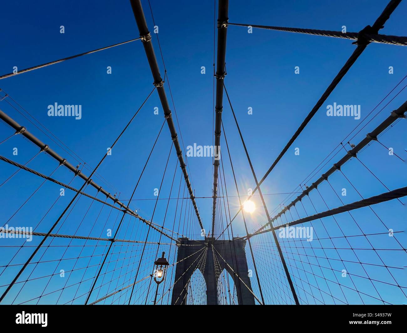 Cavi di sospensione e torre di supporto del ponte di Brooklyn con una silhouette che si affaccia su un sole luminoso e un cielo blu brillante - Immagine stock catturata con smartphone