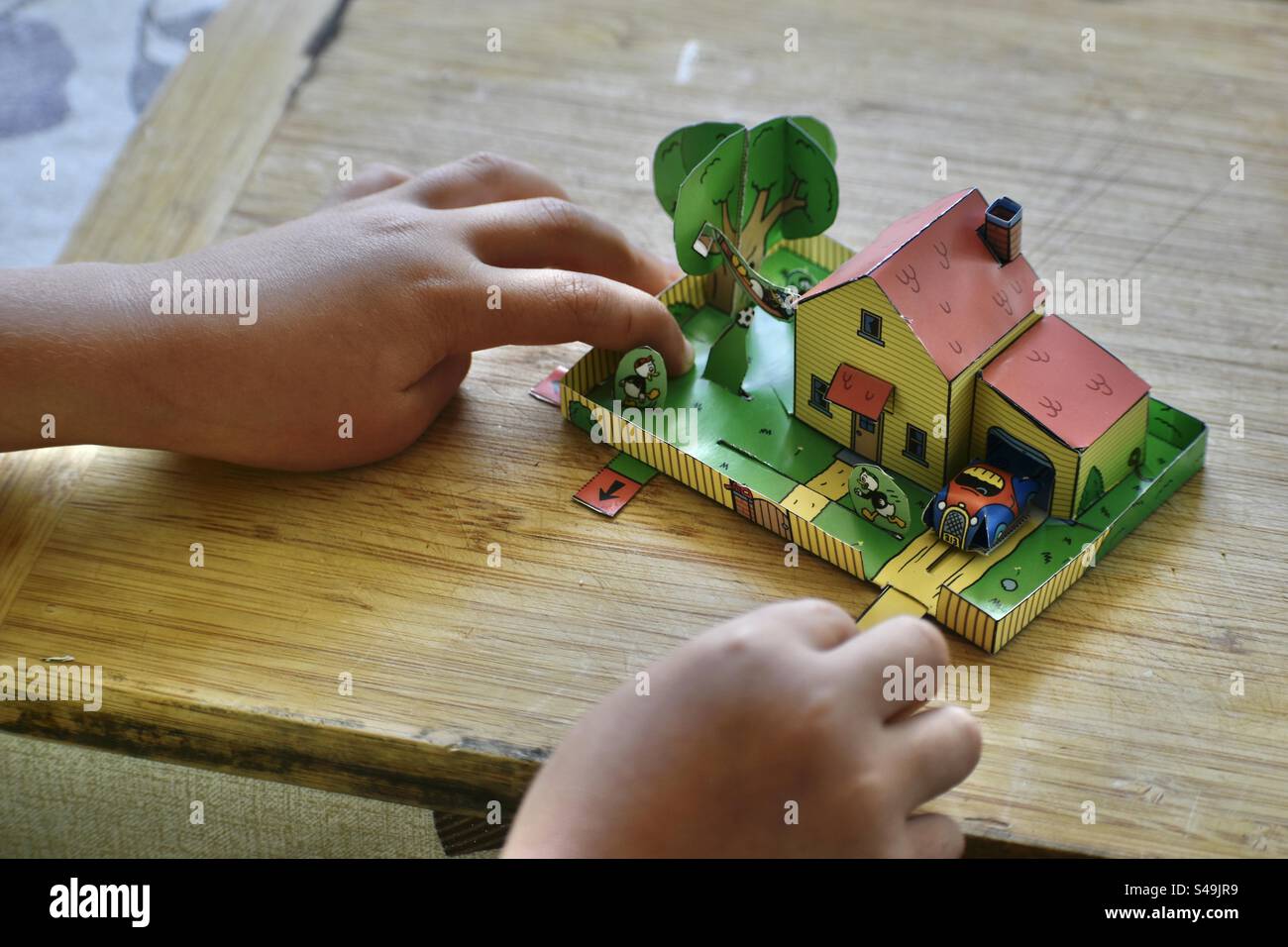 Le mani di una bambina giocano con un giocattolo ritagliato di carta o di cartone. Una casa animata con cortile o giardino. Foto Stock