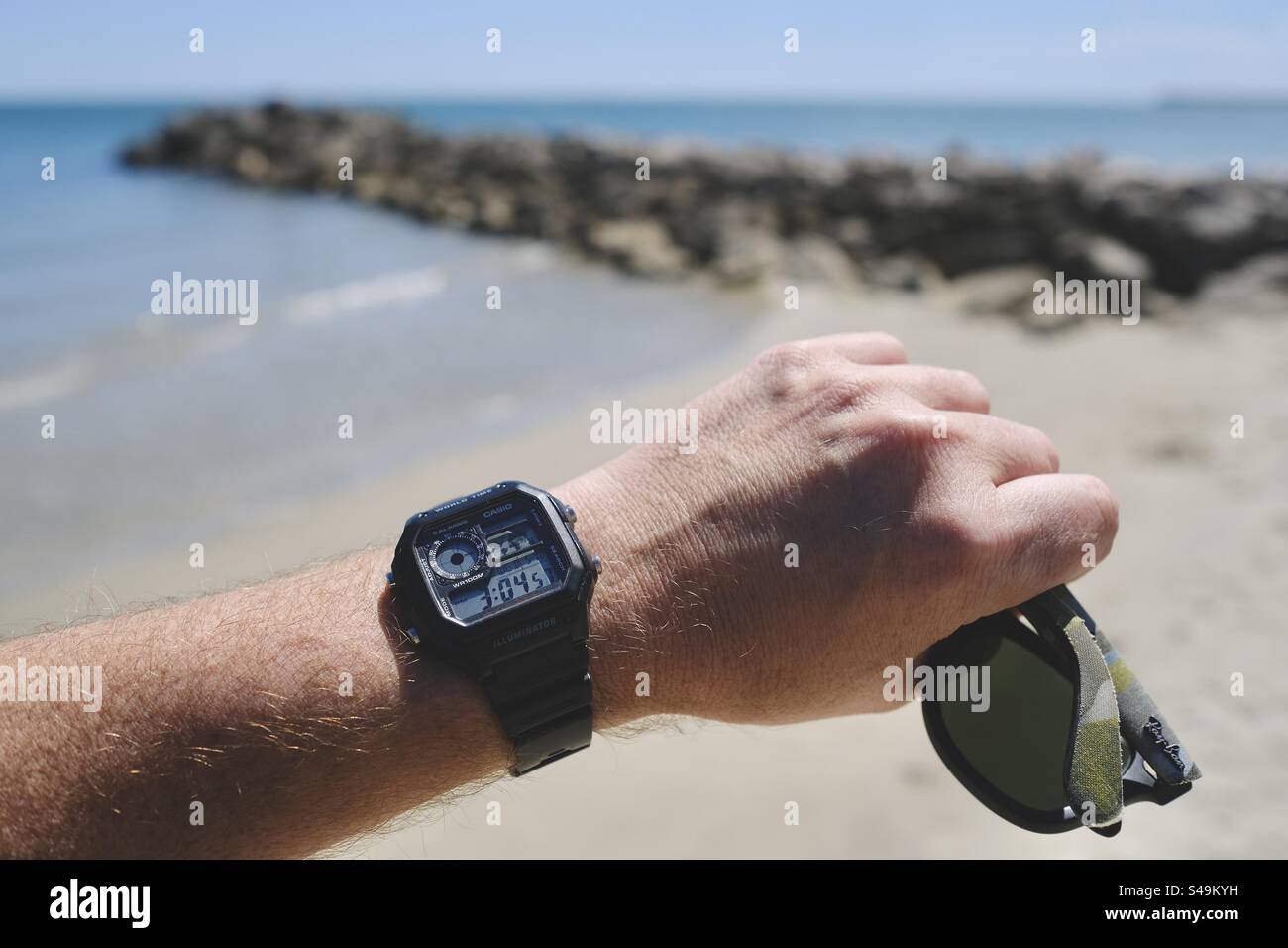 Mano di un uomo che regge un paio di RayBans camo, indossando un orologio analogico digitale Casio Royale sullo sfondo di una spiaggia e di un molo roccioso a Palavas-les-Flots, Occitanie, Francia. Foto Stock