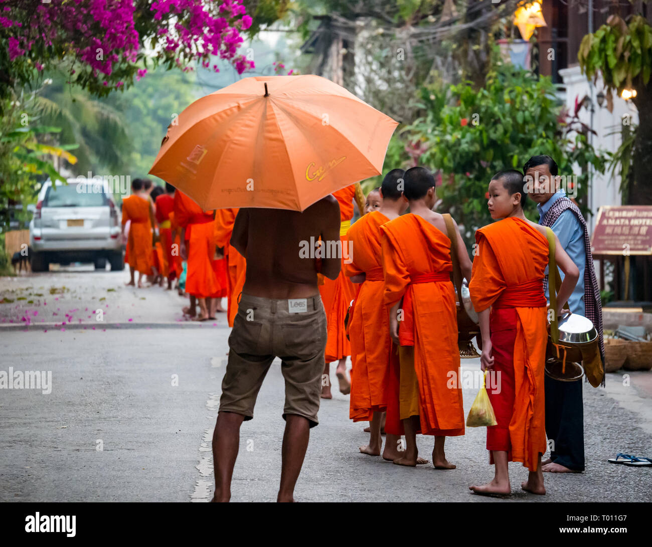 L'uomo nero con ombrello arancione guarda i monaci buddisti in abiti arancioni in coda per la cerimonia di consegna delle elemosine mattutine, Luang Prabang, Laos Foto Stock