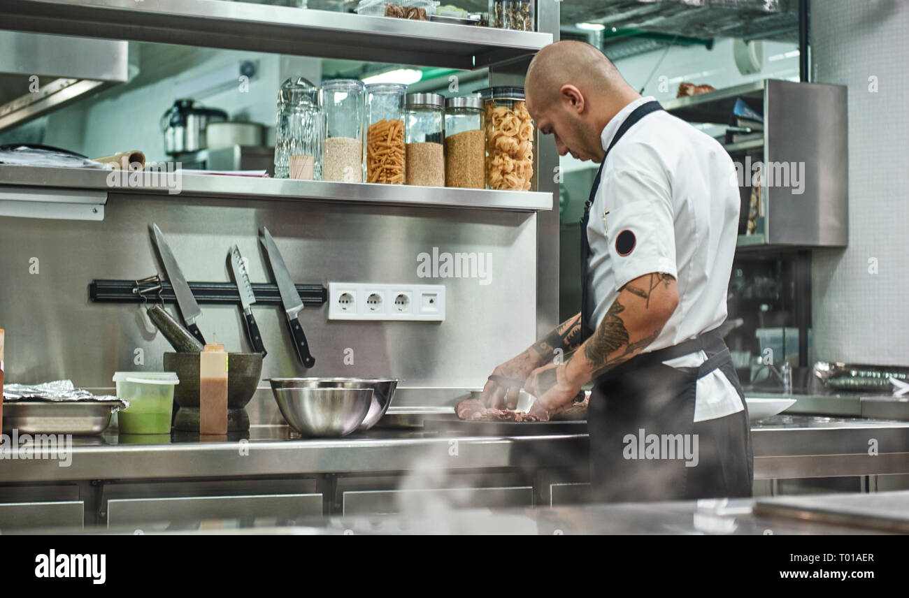 Il processo di lavorazione. Vista posteriore dei maschi di chef in grembiule e con tatuaggi sulle sue braccia il taglio di carne mentre stanno in piedi in un ristorante di cucina. Concetto culinario Foto Stock