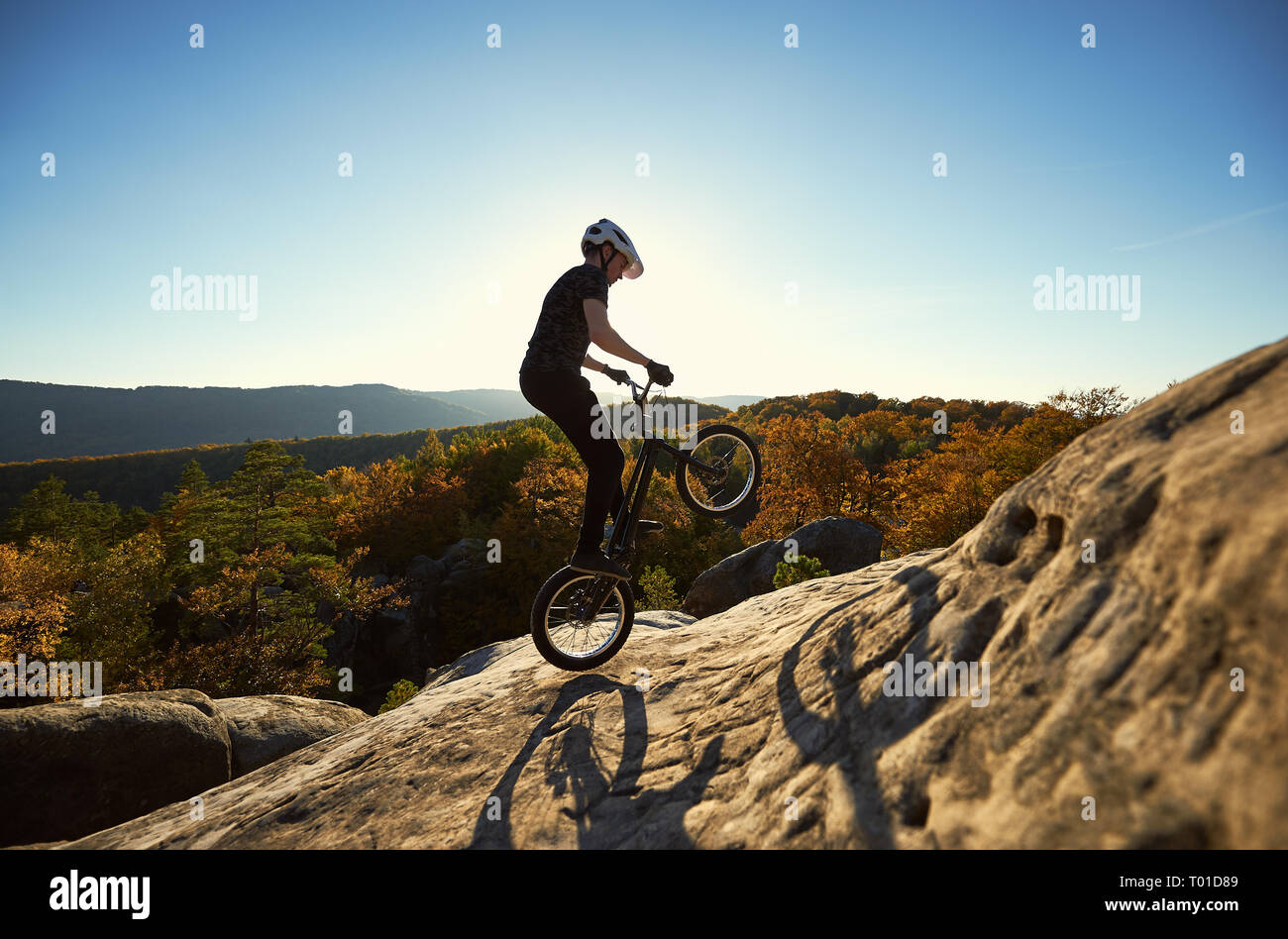Professionista sportivo ciclista salto sulla bicicletta di prova sulla parte superiore del grosso masso. Biker maschio compiendo acrobazie stunt in serata estiva, cielo blu e sunse Foto Stock