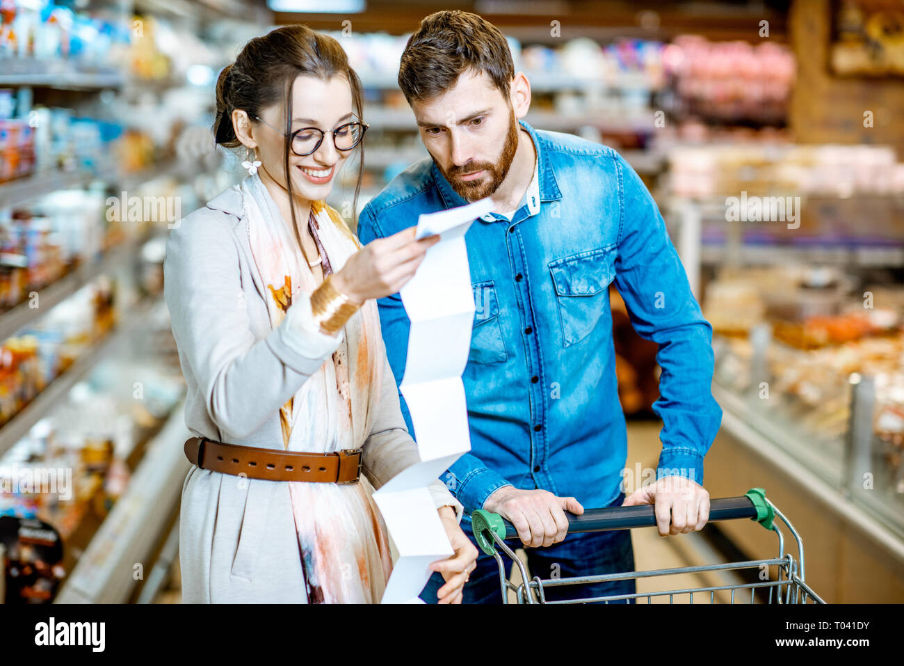 Coppia giovane comprare generi alimentari in piedi con il carrello e una lunga lista di shopping nel supermercato Foto Stock