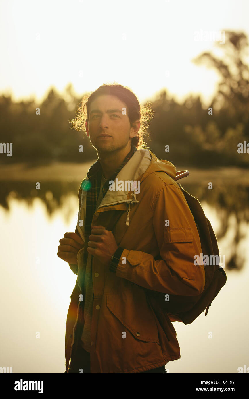 Uomo che indossa una giacca e uno zaino in piedi all'aperto accanto a un lago. viaggiatore in piedi all'aperto guardando lontano con il sole sullo sfondo. Foto Stock