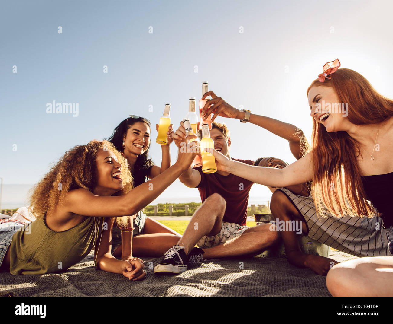 Un gruppo di giovani che partying con birre all'esterno. Multirazziale uomini e donne seduti insieme e birre di tostatura. Foto Stock