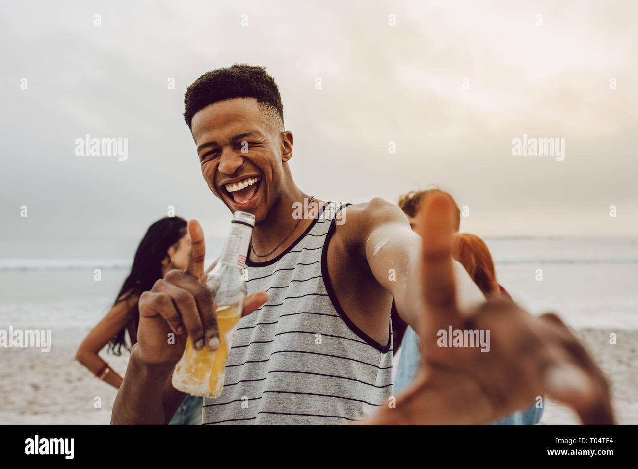 Entusiasta del giovane africano con la birra sulla spiaggia con gli amici all'indietro. Sorridente maschio con un gruppo di amici in background in spiaggia. Foto Stock