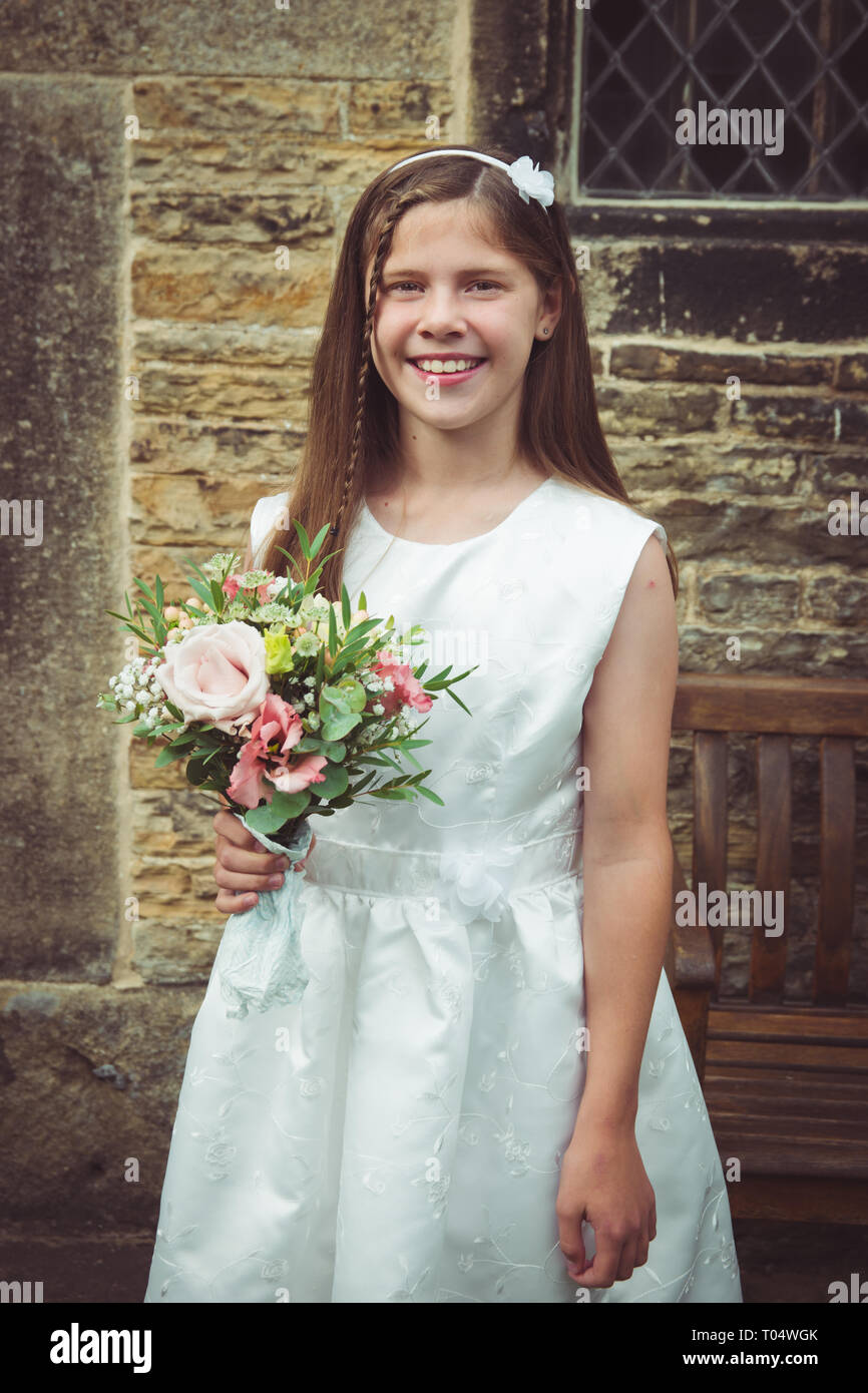 Bella sorridente tween o teen giovane damigella tenendo un rustico bouquet di fiori selvaggi, indossando un abito bianco, nella parte anteriore di un'annata chiesa di pietra Foto Stock