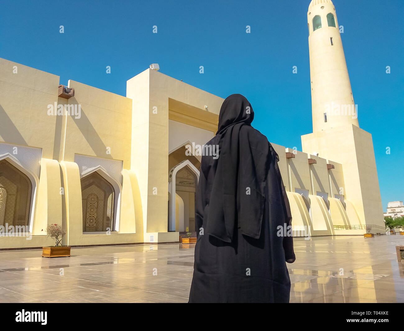 Retro della donna con abito abaya guardando la facciata e il minareto alto stato di grande moschea a Doha, in Qatar, il Medio Oriente e penisola arabica. Giornata di sole Foto Stock