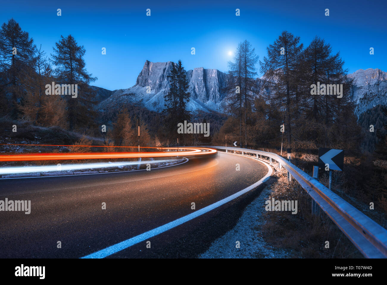 Sfocata fari auto sulla tortuosa strada di notte in autunno. paesaggio con strada asfaltata, sentieri di luce, montagne, alberi e cielo blu con la luna al crepuscolo Foto Stock