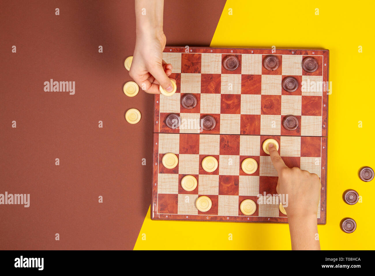 Bambino mani giocando a Dama su checker board game over giallo e blu di sfondo, vista dall'alto Foto Stock