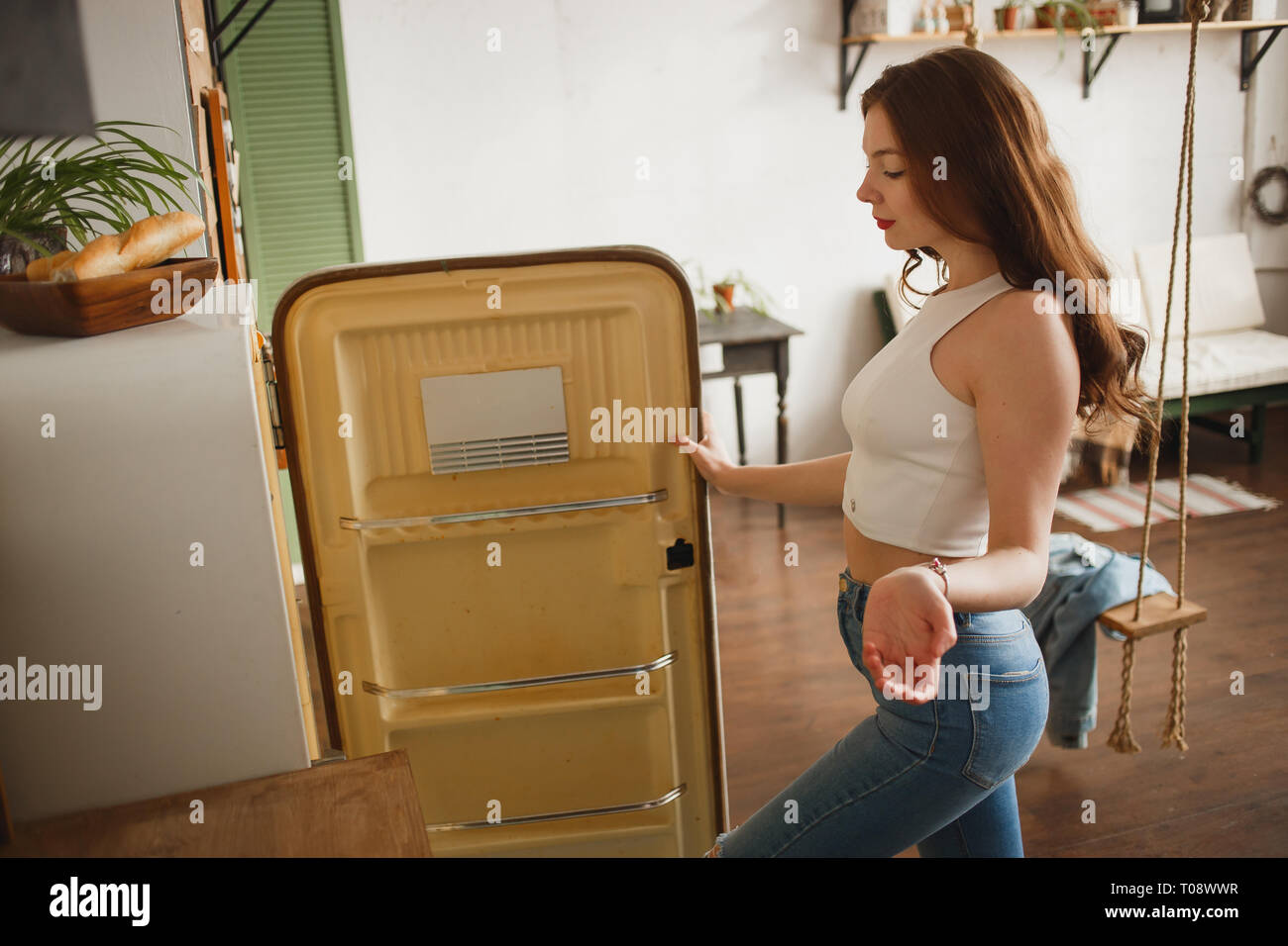 Snella ragazza in jeans aprendo la porta di vuoto retrò frigorifero e in modo sorprendente cercando in frigo. Foto Stock