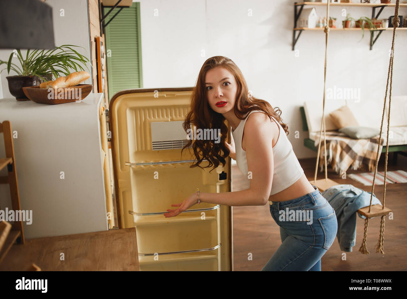 Snella ragazza in jeans aprendo la porta di vuoto retrò frigorifero e in modo sorprendente guardando nella telecamera. Foto Stock