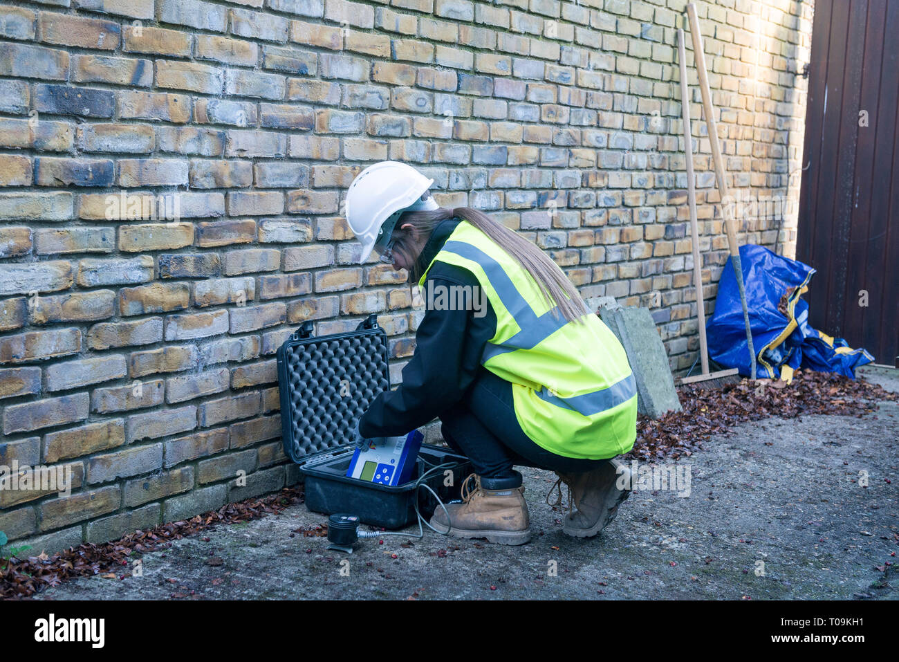 Una femmina di geometra ambientale utilizza attrezzature specialistiche mentre in alta viz, elmetto e ingranaggio di sicurezza per ispezionare un edificio sito / Proprietà commerciale Foto Stock