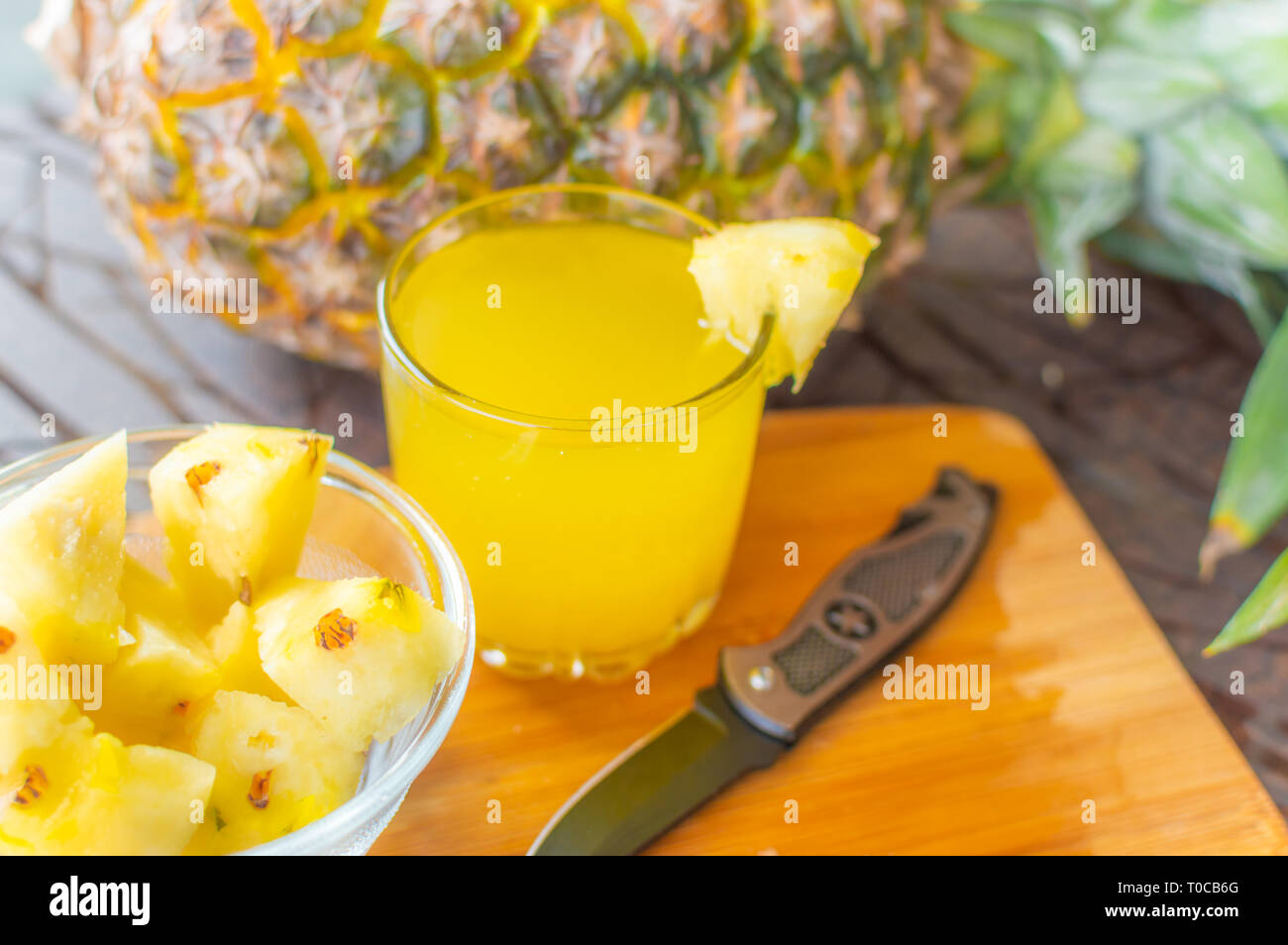 Vista laterale di ananas intero mantenuta su un tavolo in legno oltre ad un bicchiere riempito con succo di ananas e una ciotola di vetro con fette di ananas e un coltello kep Foto Stock